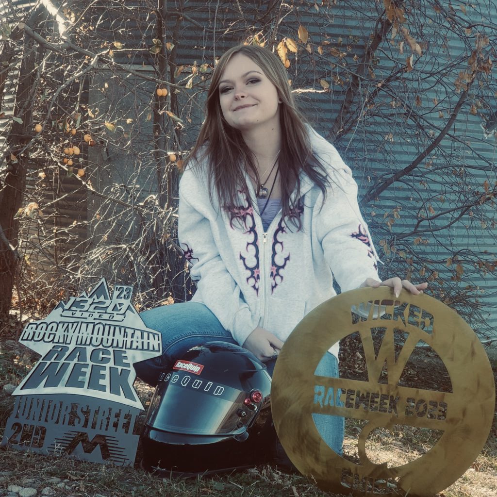 A smiling young woman in a hoodie sits beside a racing helmet and trophies, with trees and a shed in the background.