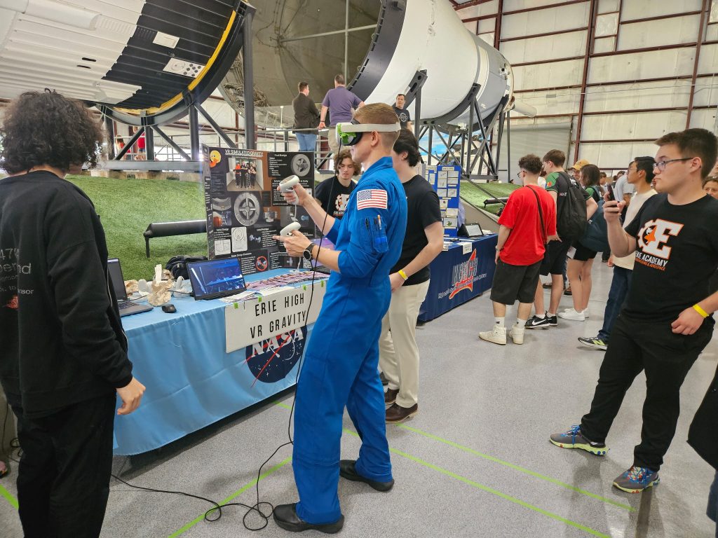 A group of students at a science fair, with one in a blue astronaut suit using a VR headset at a booth.