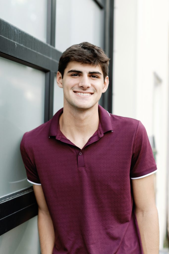 A young man with short dark hair smiles while leaning against a glass window, wearing a burgundy polo shirt.