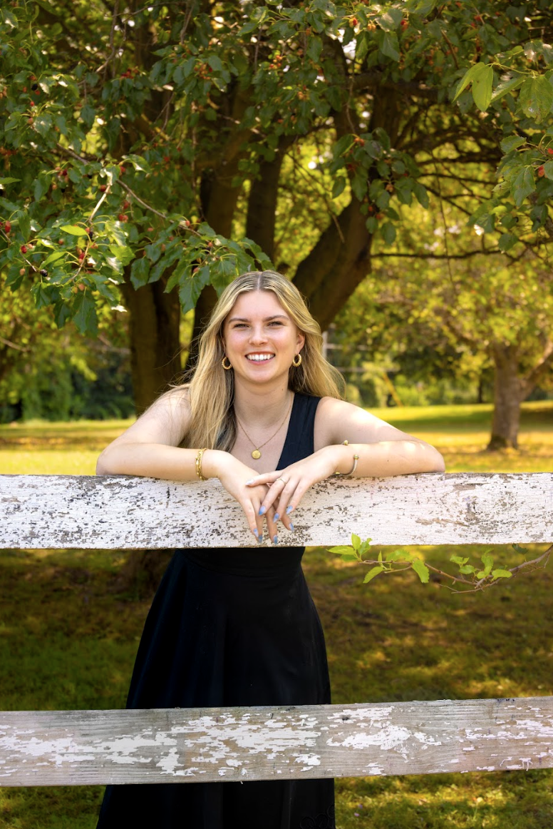 A smiling young woman in a black dress leans on a weathered wooden fence, surrounded by lush greenery.