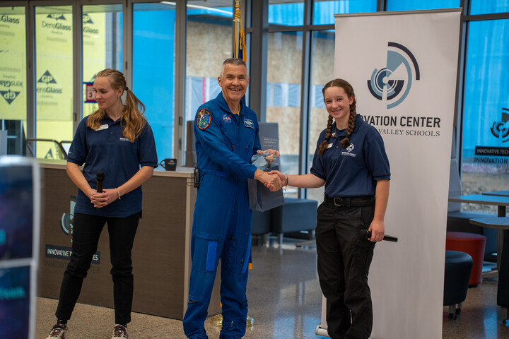 Two students in blue shirts stand beside Duane Carey in a blue flight suit, shaking hands at an event in a modern space.