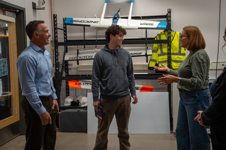 Three people are engaged in conversation in a workspace, with equipment and safety gear visible in the background.