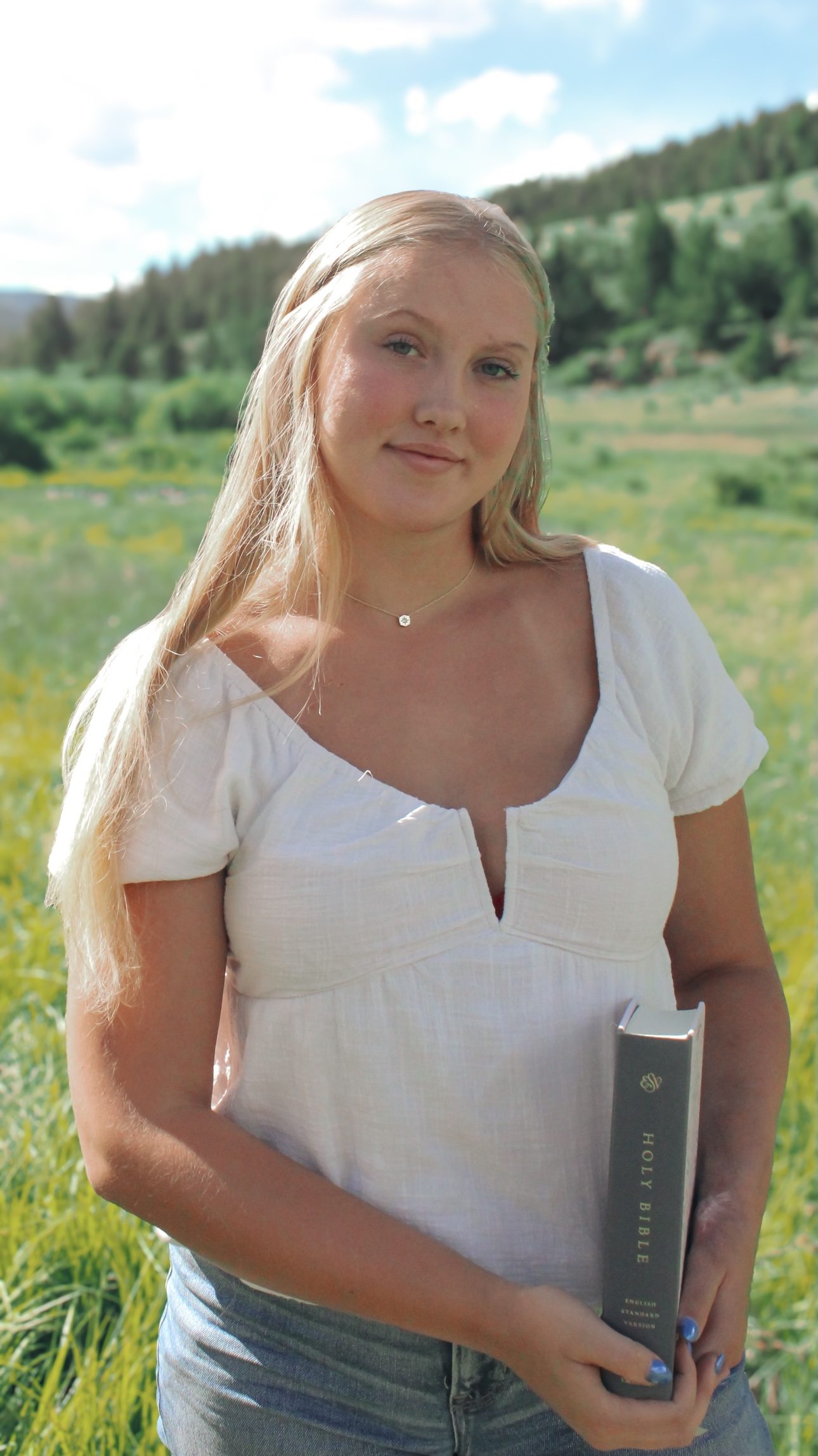 A young woman in a white top holds a Bible in a sunny green field with mountains in the background.
