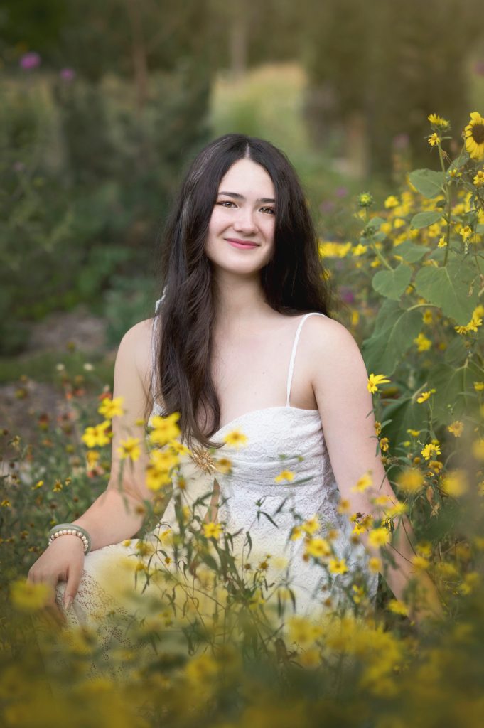 A young woman sits among yellow flowers, wearing a white dress and smiling softly in a natural setting.