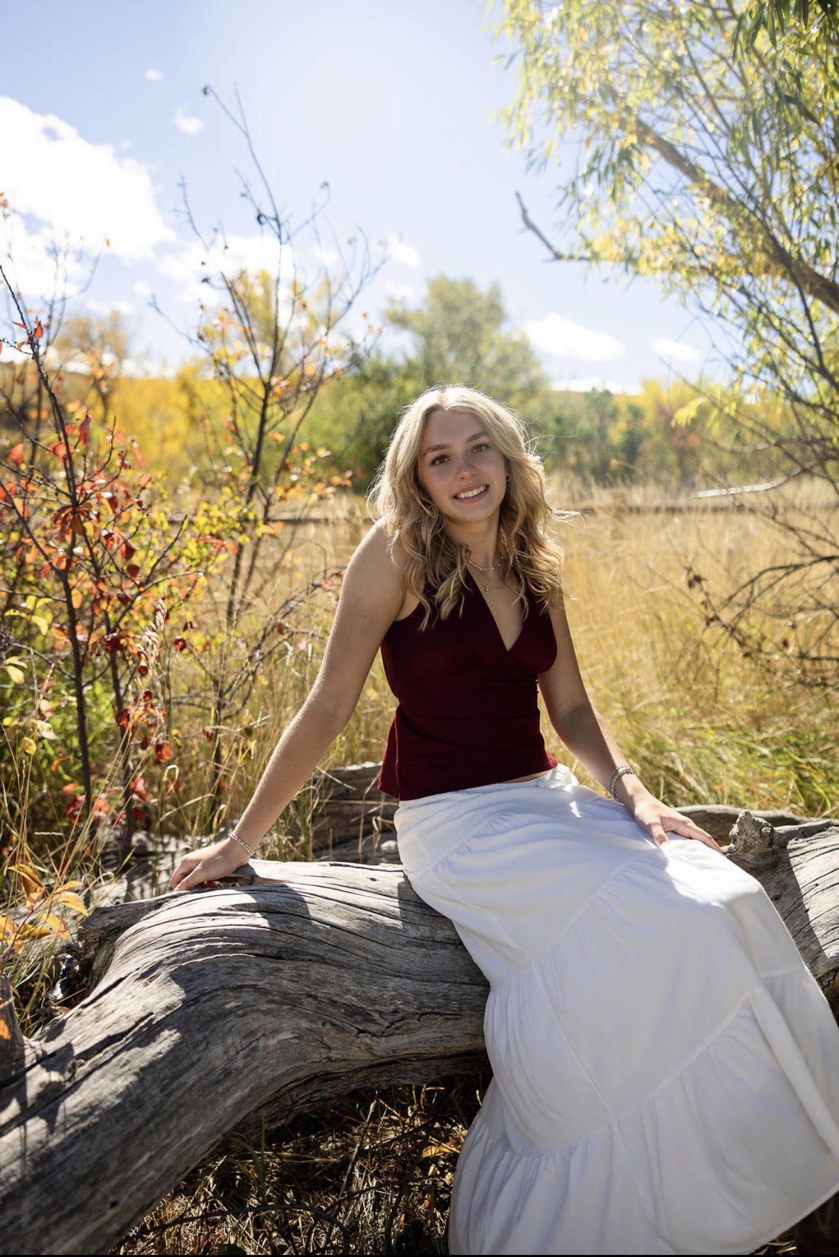 A young woman sits on a log in a sunny field, wearing a burgundy top and a white skirt, surrounded by autumn foliage.
