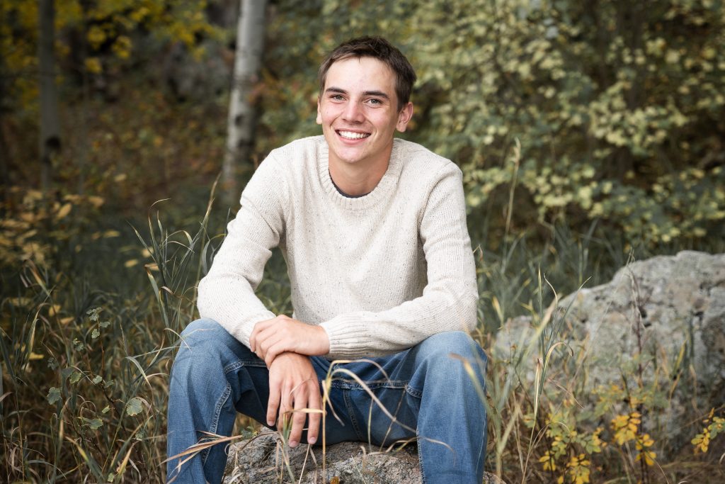A young man smiles while sitting on a rock, surrounded by tall grass and trees with autumn leaves.
