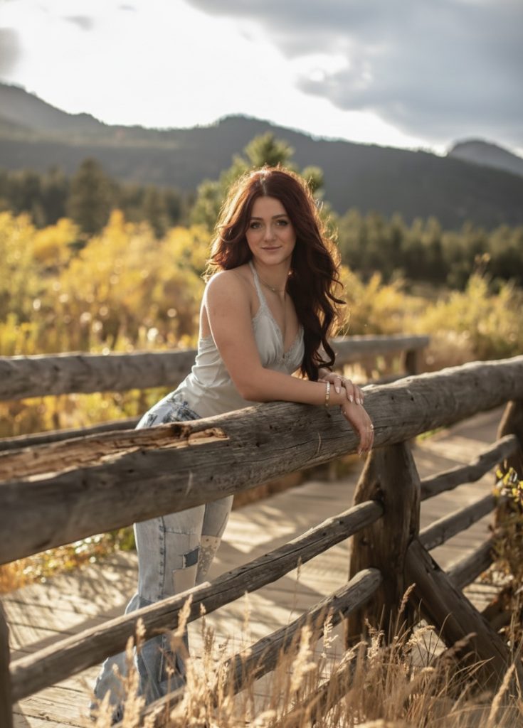 A woman with long red hair leans on a wooden fence, surrounded by autumn foliage and mountains in the background.