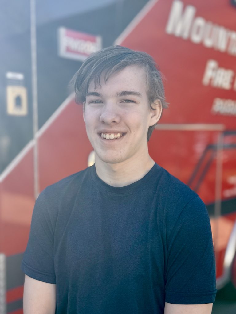 A young person with light brown hair smiles in front of a red and black fire truck.