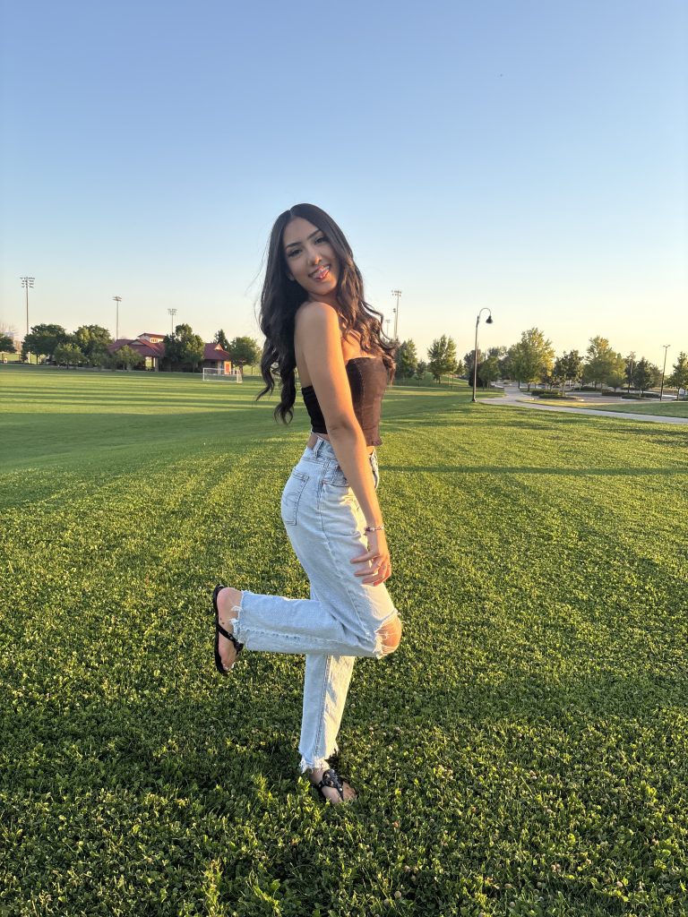 A woman poses playfully on a grassy field, wearing a black top and light blue ripped jeans, smiling at the camera.