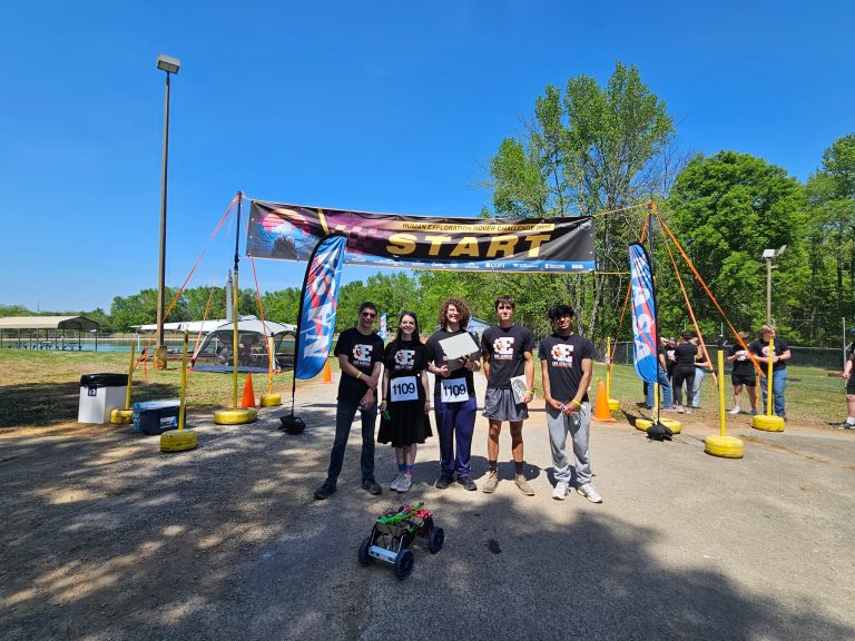 A team of six stands at the start line of a rover challenge, holding a model rover under a clear blue sky.