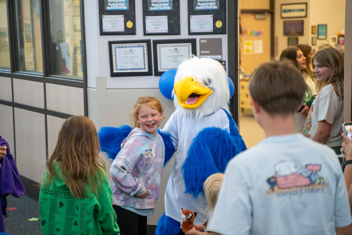 A blue and white eagle mascot interacts cheerfully with children in a school hallway decorated with certificates.