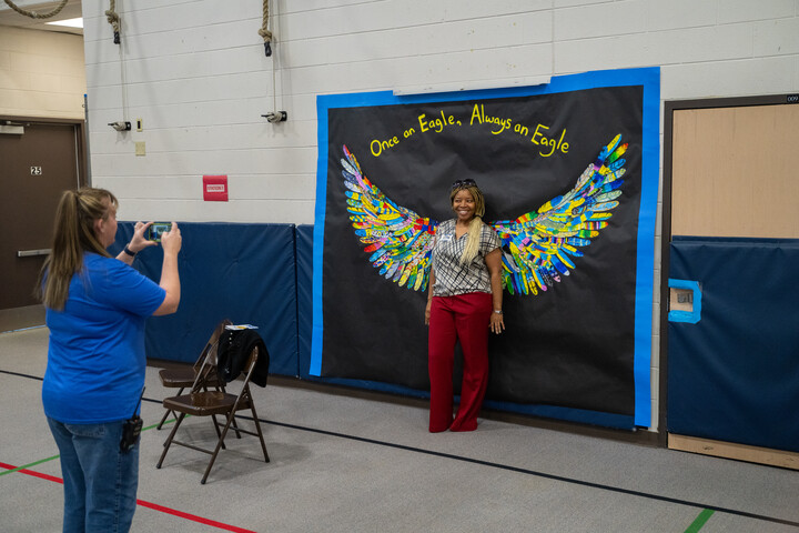 A woman poses in front of a colorful wing mural while another person takes her photo in a gym setting.