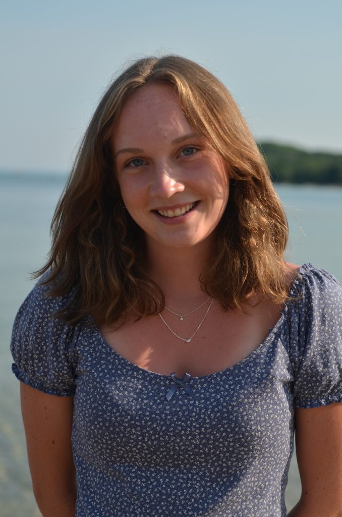 A young woman with wavy hair smiles at the camera, wearing a floral dress, with a serene lake in the background.