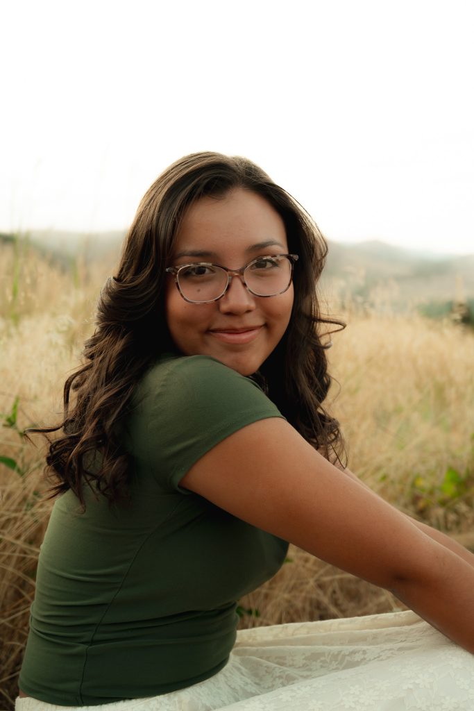 A young woman with long, wavy hair sits in a field, wearing glasses and a green shirt, smiling at the camera.