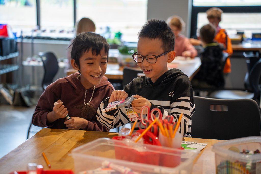 Two children are smiling and working together on a creative project with art supplies on a table.