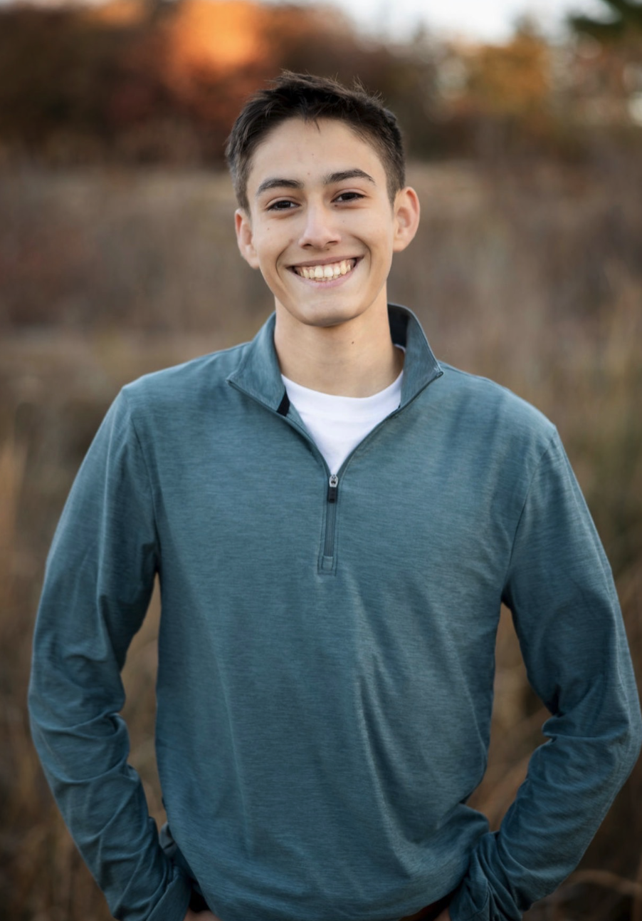 Smiling young man in a teal pullover stands outdoors, with nature blurred in the background.