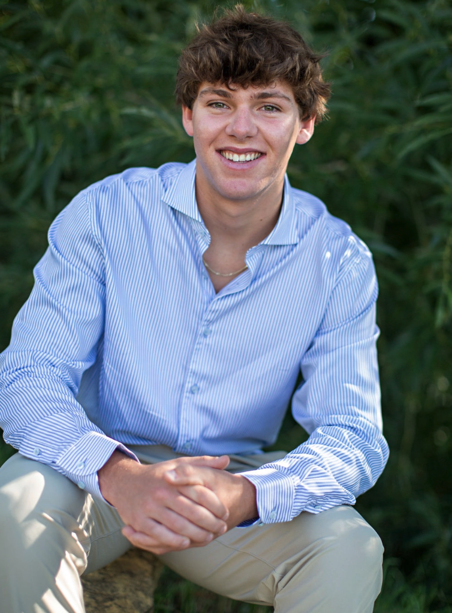 Young man sitting outdoors, smiling, wearing a striped blue shirt and light pants, with greenery in the background.