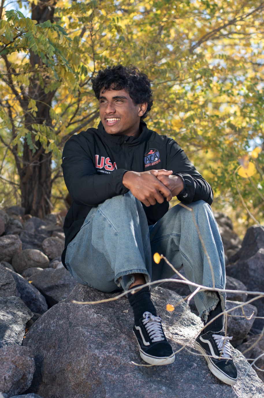 A young man sits on large rocks, smiling, wearing a black jacket and denim jeans, surrounded by autumn foliage.