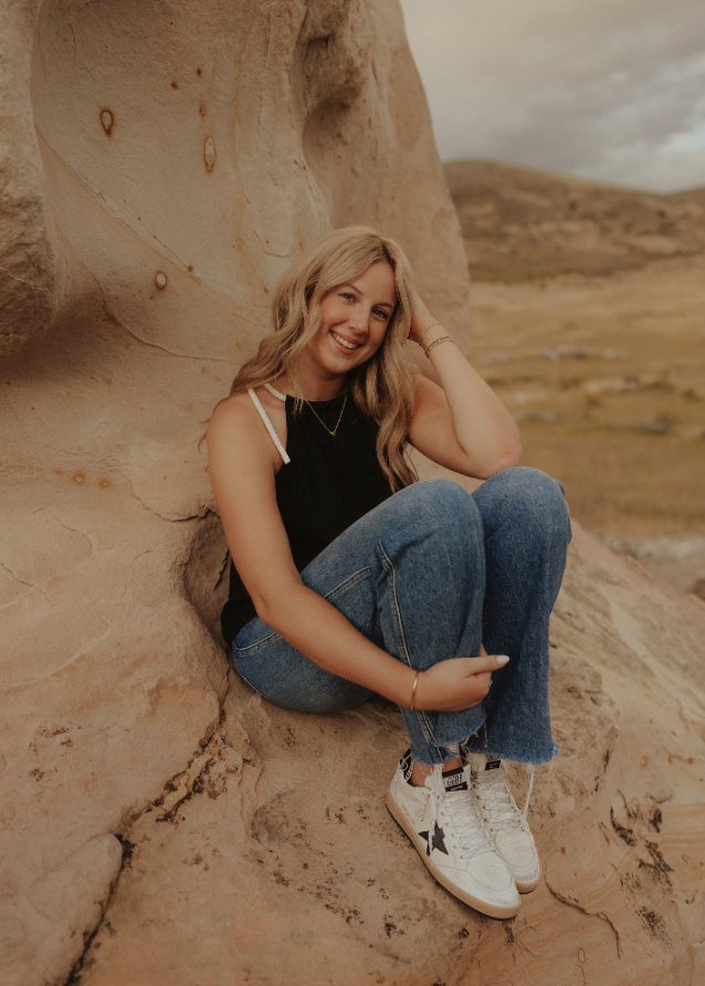 A young woman sits on a rock, wearing a black top and blue jeans, smiling with a scenic landscape in the background.