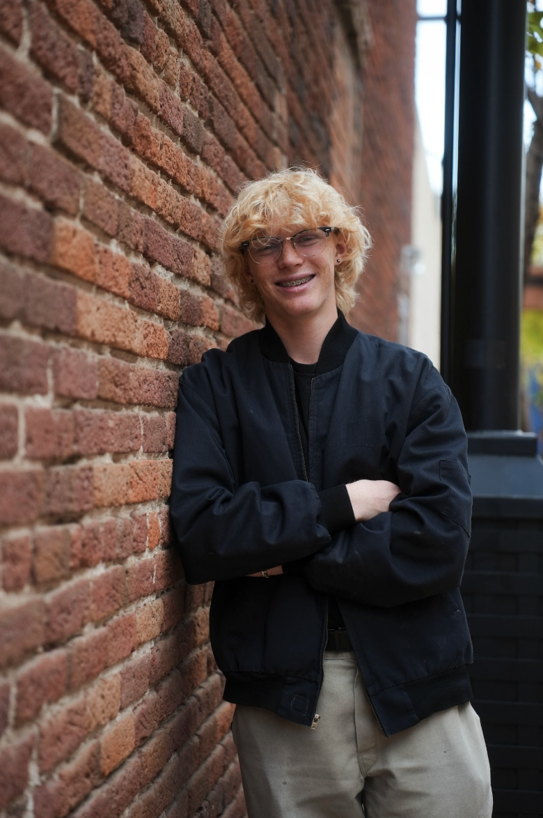 A young person with curly blonde hair and glasses stands against a brick wall, smiling with arms crossed.