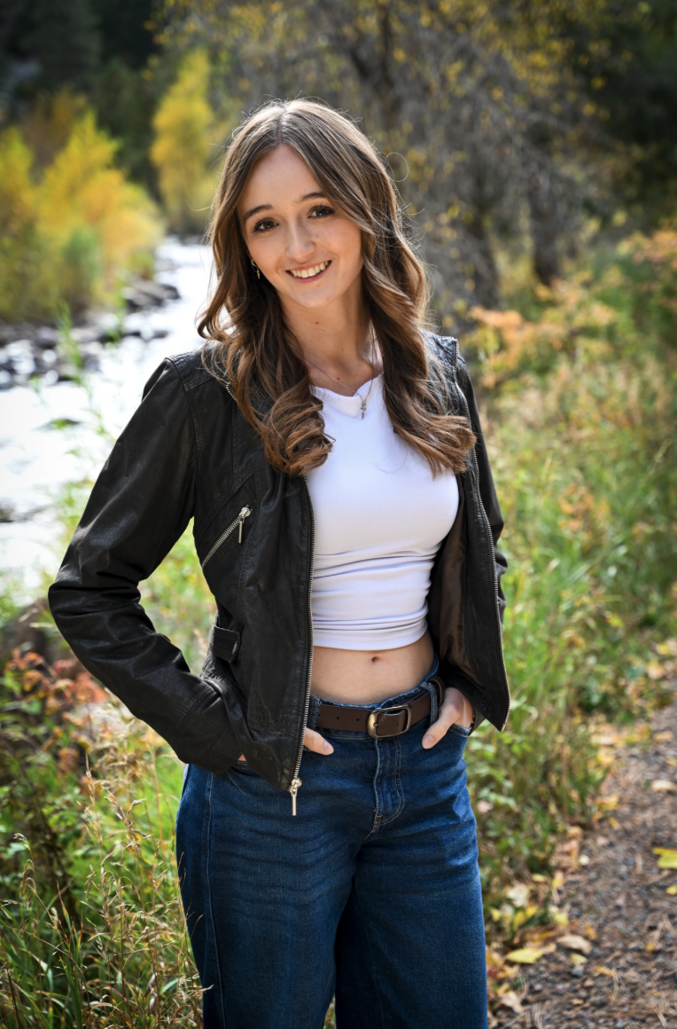 A young woman stands by a river, wearing a black jacket and blue jeans, smiling amidst colorful autumn foliage.