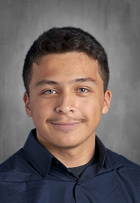 A young man with short dark hair, wearing a navy blue shirt, smiles at the camera against a gray background.