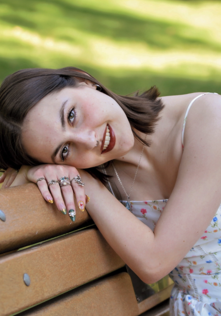A young woman rests her head on a wooden bench, smiling, with floral attire and colorful nail art.