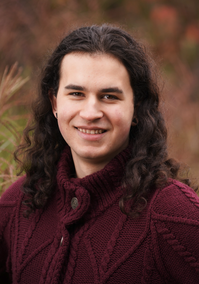 A young person with long curly hair smiles, wearing a maroon sweater, against a blurred natural background.