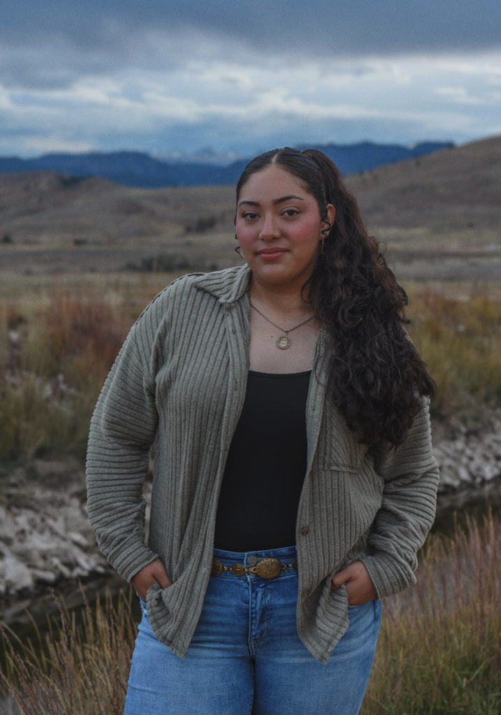 A young woman stands outdoors in a green cardigan and jeans, with a scenic landscape and mountains in the background.