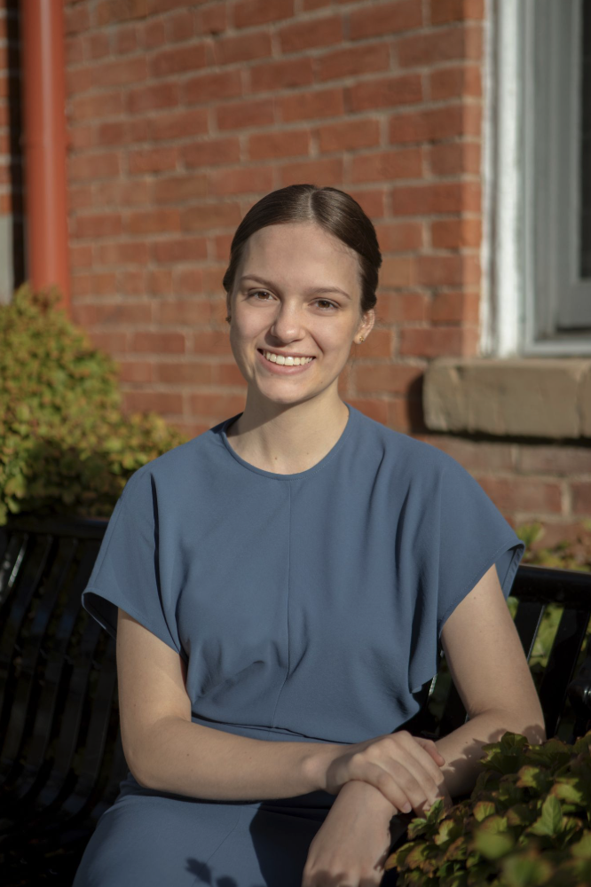 A woman with a warm smile sits on a bench, wearing a blue dress, with a brick wall and greenery in the background.