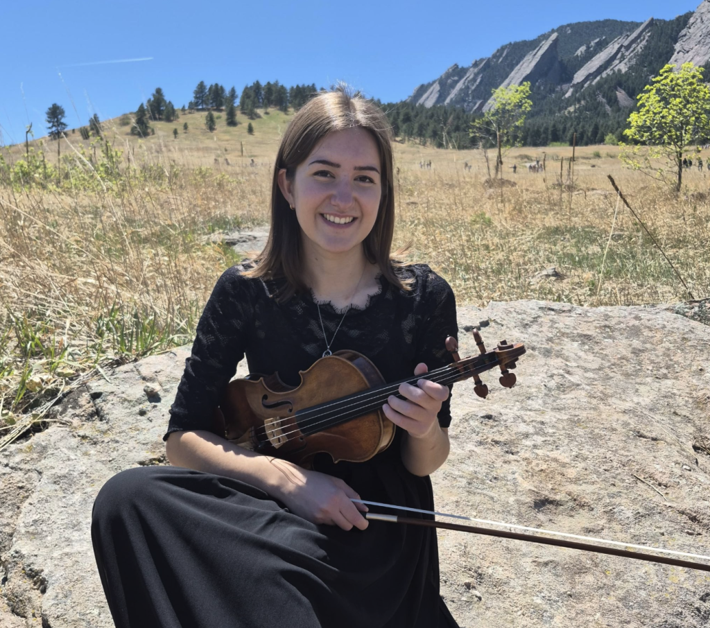 A young woman in a black dress sits on a rock, holding a violin with a scenic mountain background.
