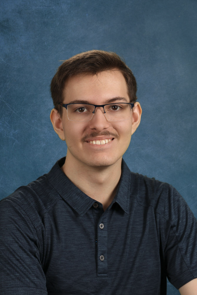 A young man with glasses and a mustache smiles, wearing a dark polo shirt against a blue background.