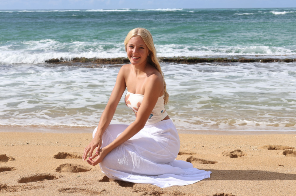 A person with long blonde hair sits on the beach, smiling, with waves and a sandy shoreline in the background.