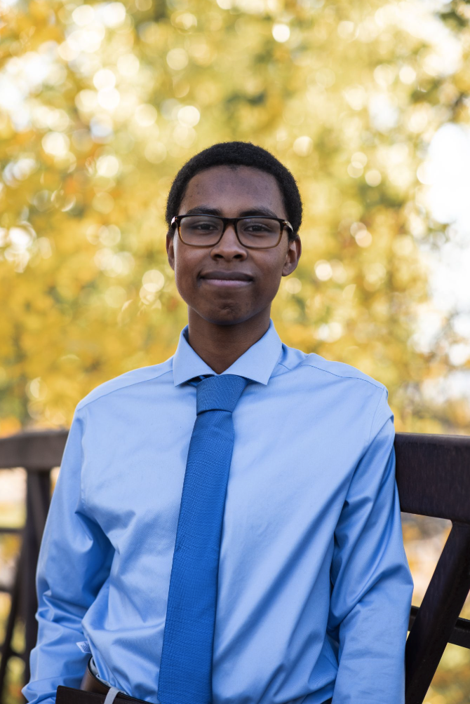 A young man in a light blue shirt and tie stands outdoors, smiling against a backdrop of blurred autumn foliage.
