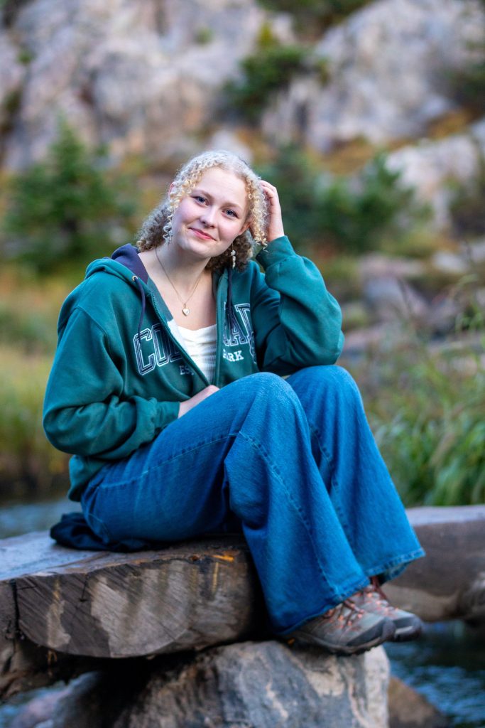A person with curly hair sits on a log by a river, wearing a green hoodie and blue jeans, smiling at the camera.