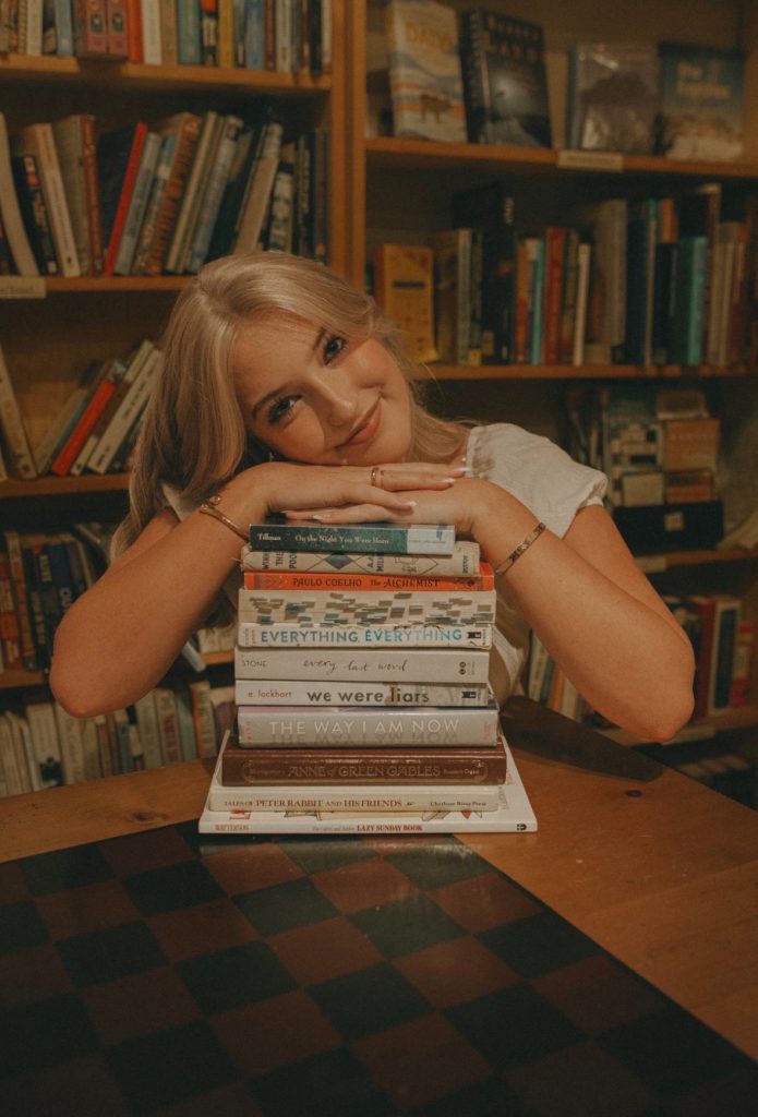 A young woman rests her chin on a stack of books in a cozy library setting, smiling at the camera.
