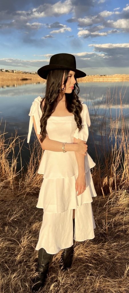 A woman in a white tiered dress and black hat stands by a reflective lake, surrounded by dry grass and blue skies.