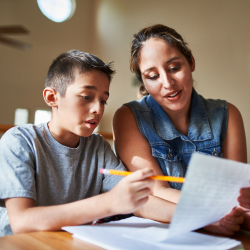A woman helps a boy with his homework at a table, both focused on a paper in front of them.