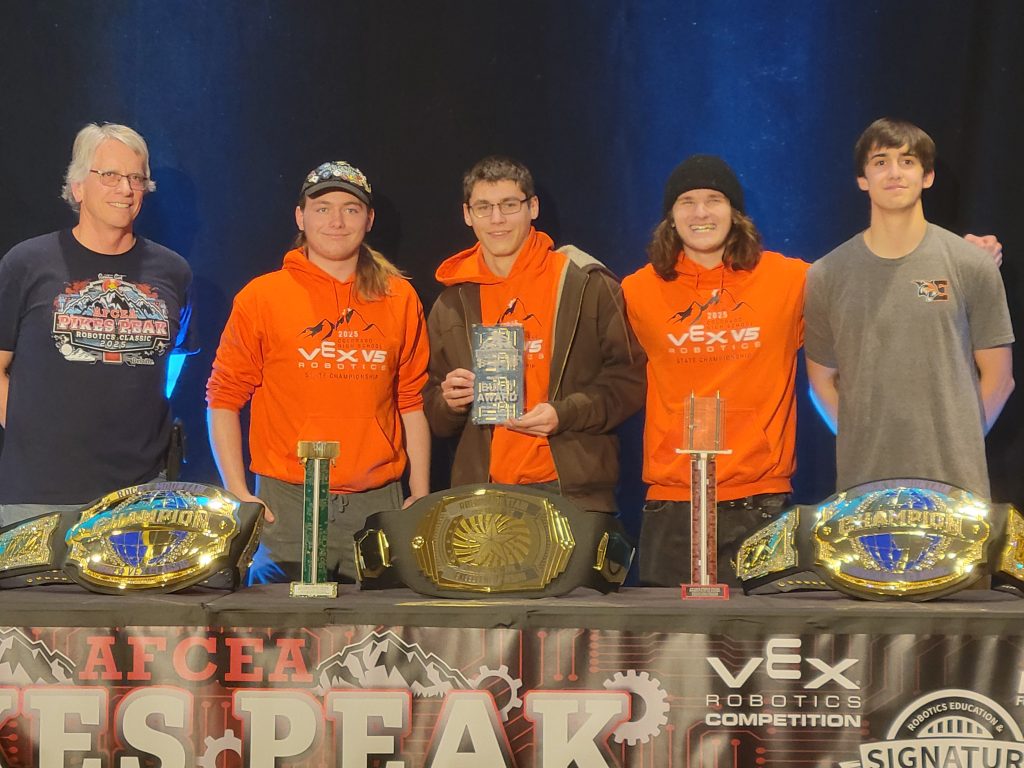 Five individuals stand behind a table displaying trophies and championship belts at a robotics competition event.