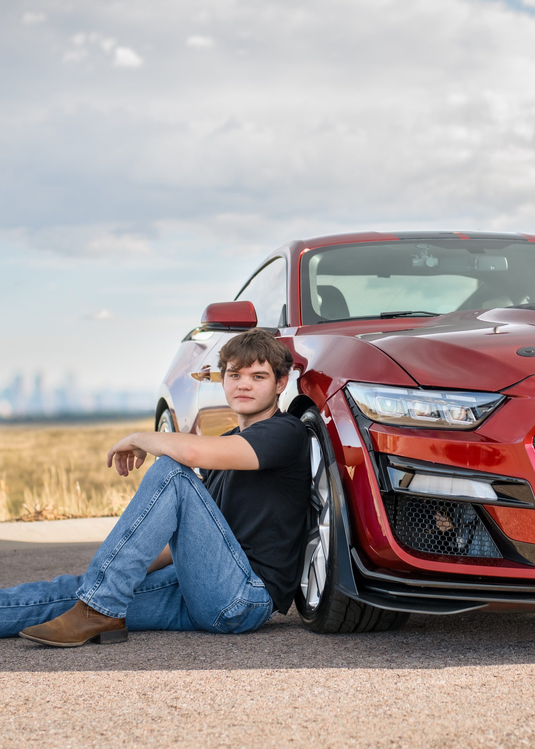 A young man sits beside a red sports car on a road, with a cloudy sky and distant cityscape in the background.