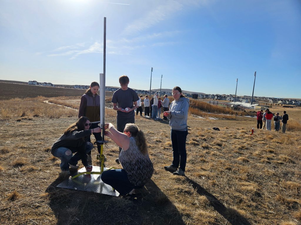 Students work together outdoors to assemble a rocket on a sunny day, with fields and houses in the background.