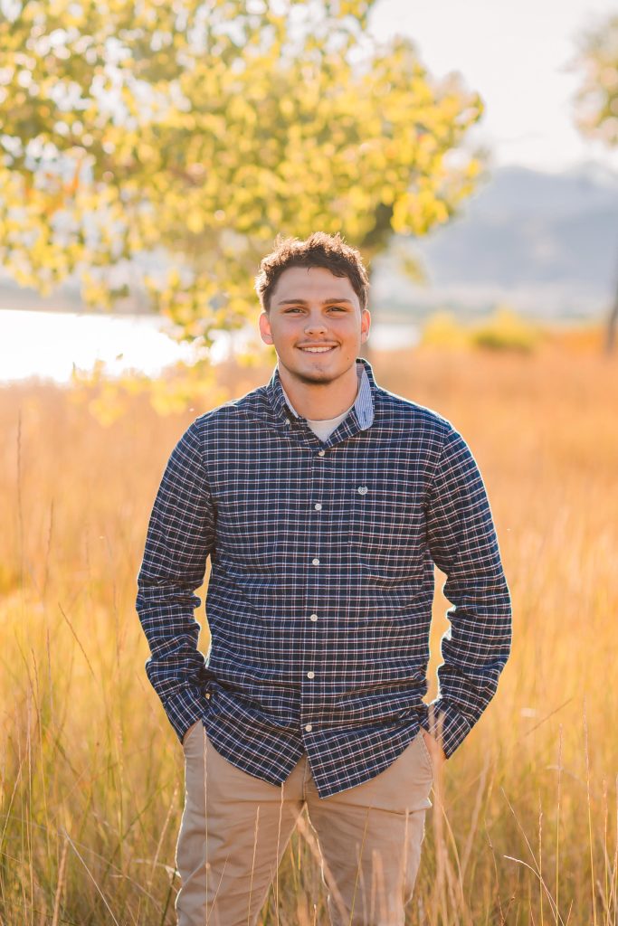 A young man stands in a field of tall grass, smiling, wearing a plaid shirt and khaki pants, with trees and water in the background.