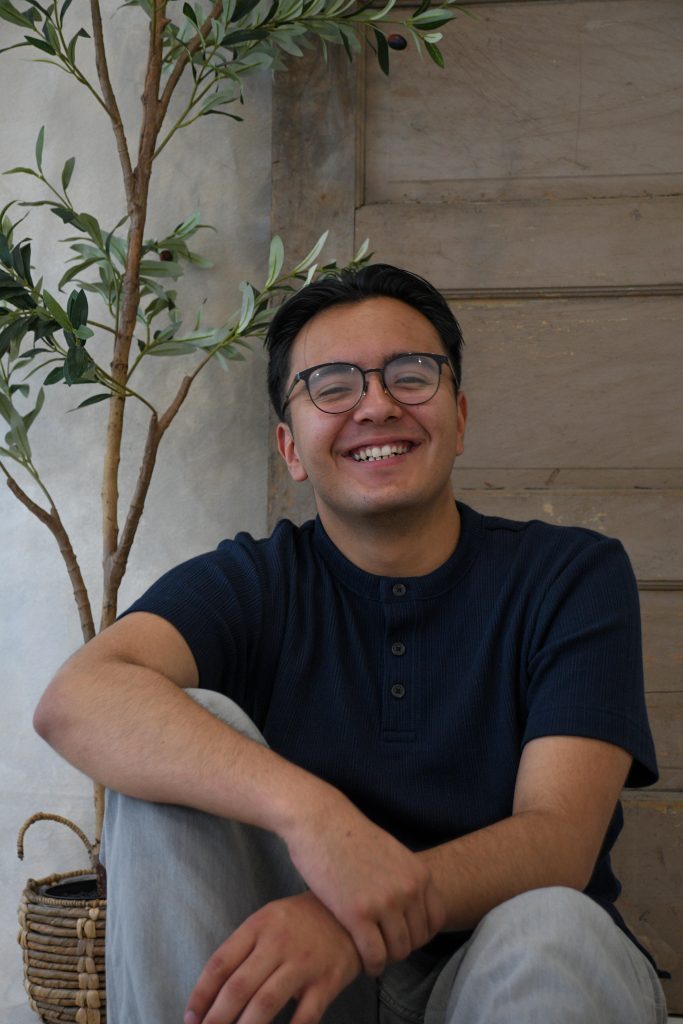 A smiling young man sits on the floor beside a potted olive tree against a wooden wall. He wears a navy shirt.