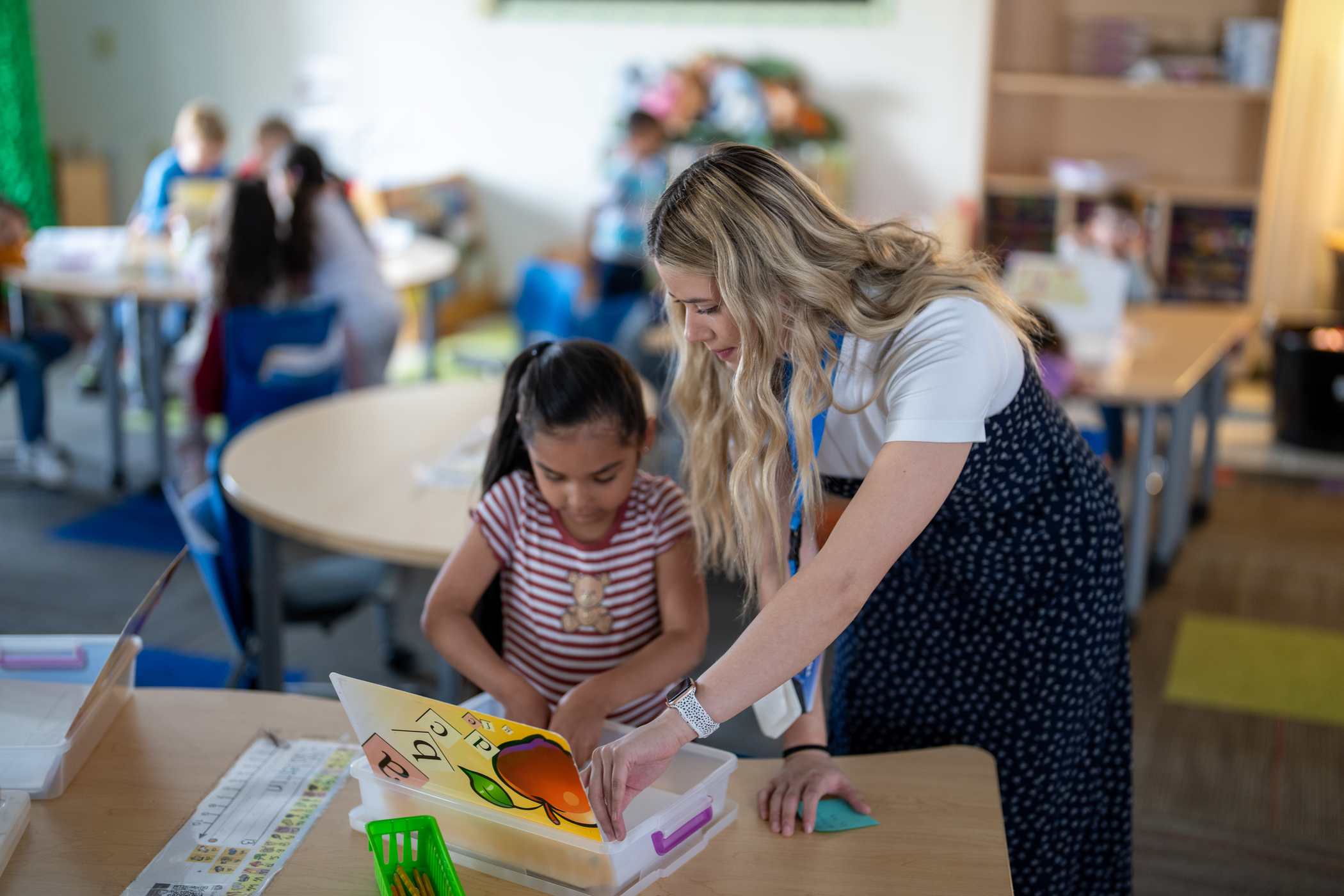 Una profesora ayuda a una niña a realizar una actividad en una mesa del aula, rodeada de otros alumnos que están aprendiendo.