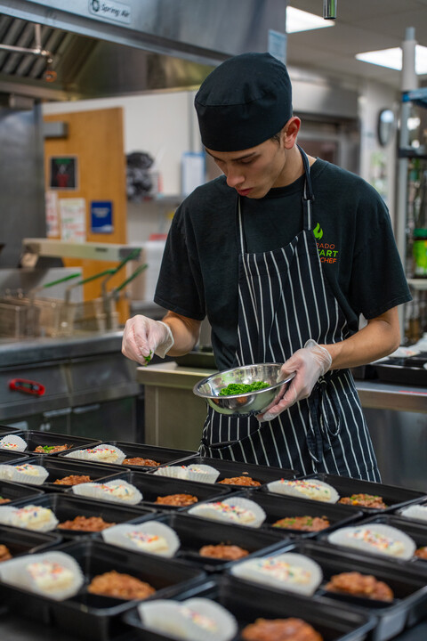 Un estudiante, en una cocina, añade guarniciones a los platos servidos en bandejas negras, mientras prepara la comida para un proyecto de colaboración con la comunidad.