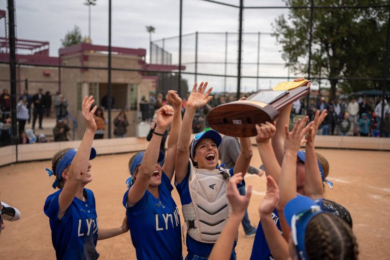 Lyons Lions softball team celebrates with the championship trophy on the field, surrounded by cheering teammates.