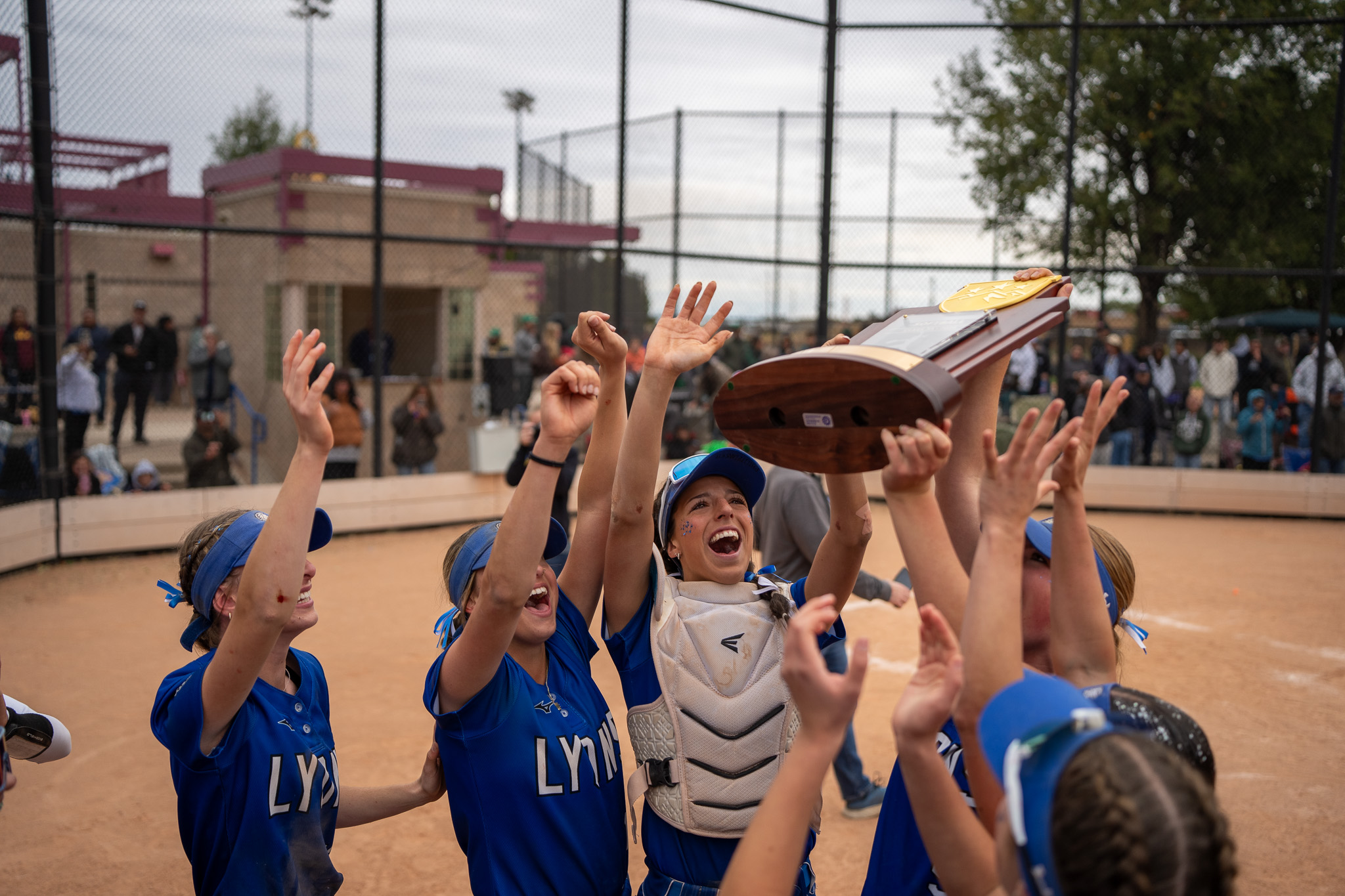 Lyons Lions softball team celebrates with the championship trophy on the field, surrounded by cheering teammates.